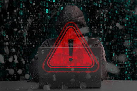A hooded hacker behind a laptop with a red glowing warning with a triangle and exclamation mark. Binary code overlays the scene, emphasizing the concept of data leakage and cybercrimeの写真素材