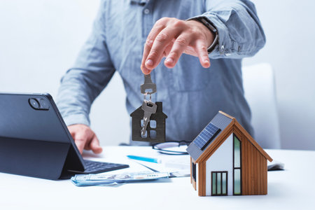 A model home with solar panels and keys in the hands of a realtor. Concepts of home selling and buying and real estate investingの写真素材