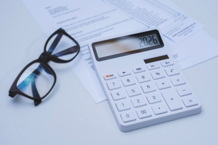 White calculator displaying the number 2026, placed on financial documents with black eyeglasses nearby. Concept business planning and tax calculationの写真素材