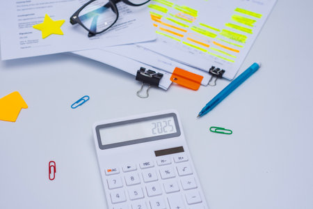 Finance and budget planning concept with eyeglasses, a calculator displaying 2025. Documents with highlighted text, and colorful office supplies on a white deskの写真素材