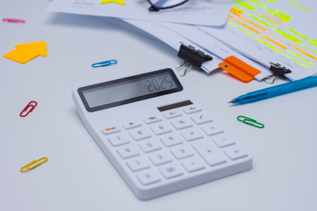 Finance and budget planning concept with eyeglasses, a calculator displaying 2025. Documents with highlighted text, and colorful office supplies on a white deskの写真素材