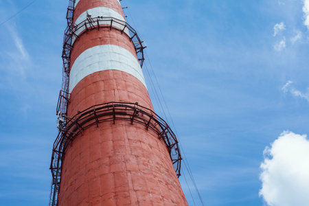 Low angle view of a tall industrial pipe with red and white stripes against a cloudy blue sky. The...