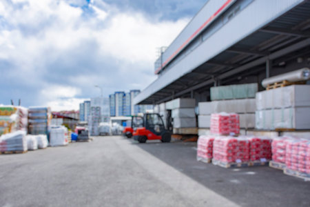 Blurred image industrial warehouse with stacks of construction materials on pallets and a red forklift. Outdoor logistics center or building supply depot with high-rise buildings in the backgroundの写真素材