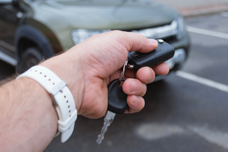 A man holds a key fob with a car key in a parking lot with a car in the background. Concept of...