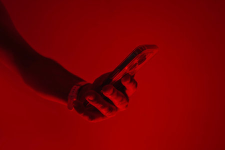 Close-up of a man's hand holding a modern smartphone under bright red studio lighting. Concept of digital cybersecurity, data privacy, hacker activity and darknetの写真素材