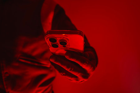Modern smartphone in a man's hand. The man holds the device under red studio light. The concept of social communication, digital cybersecurity, hacker activity and darknetの写真素材