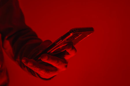 Close-up of a man's hand holding a modern smartphone under bright red studio lighting. Concept of digital cybersecurity, data privacy, hacker activity and darknetの写真素材