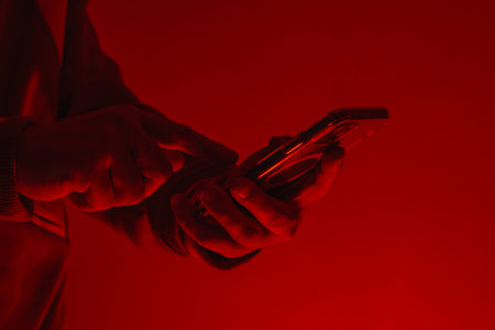 Close-up of a man's hand holding a modern smartphone under bright red studio lighting. Concept of...