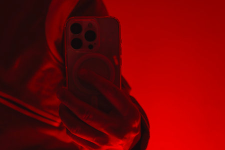 Modern smartphone in a man's hand. The man holds the device under red studio light. The concept of social communication, digital cybersecurity, hacker activity and darknetの写真素材