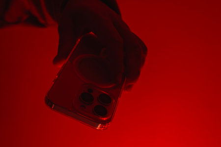 Modern smartphone in a man's hand. The man holds the device under red studio light. The concept of social communication, digital cybersecurity, hacker activity and darknetの写真素材
