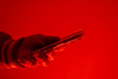 Modern smartphone in a man's hand. The man holds the device under red studio light. The concept of social communication, digital cybersecurity, hacker activity and darknetの写真素材