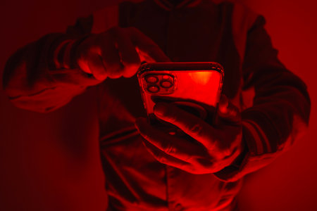 Close-up of a man's hand holding a modern smartphone under bright red studio lighting. Concept of digital cybersecurity, data privacy, hacker activity and darknetの写真素材