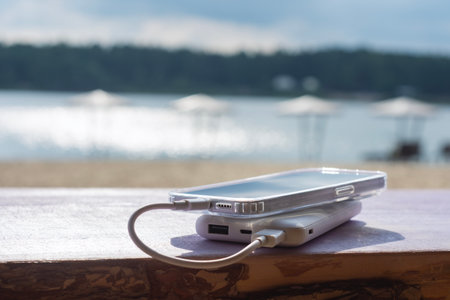 Modern smartphone is visibly charging from a power bank on a wooden table with a blurred backdrop of a sandy beach and parasols. Concept portable technology for leisure and travelの写真素材