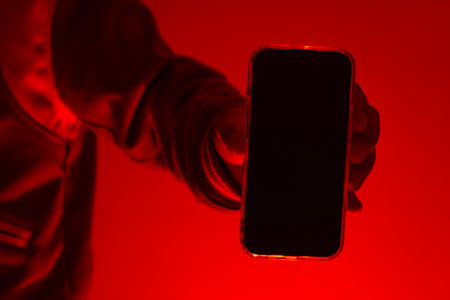 Modern smartphone in a man's hand with a close-up mockup of the screen. The man holds the device under red studio light. Concept of digital cybersecurity, hacker activity and darknetの写真素材