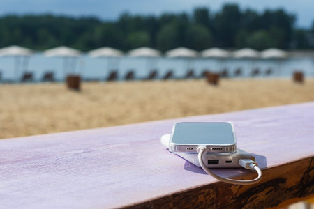 Modern smartphone is visibly charging from a power bank on a wooden table with a blurred backdrop of a sandy beach and parasols. Concept portable technology for leisure and travelの写真素材