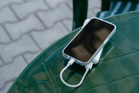 Modern smartphone connected to portable power bank via white charging cable on a green table. Mobile device for travel and emergency power solutionsの写真素材