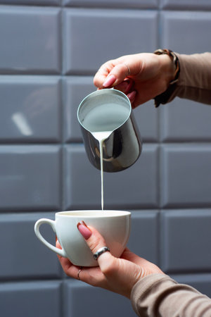 Woman hands pouring creamy milk from a stainless steel pitcher into a white coffee cup on saucer. Professional barista technique for coffee preparationの写真素材