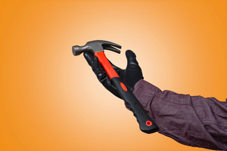 Close-up of a construction worker black gloved hand holding a claw hammer with a black and red handle against a orange backgroundの写真素材