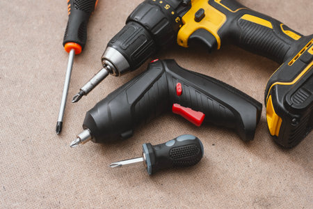 Set power tools including yellow cordless drill with battery pack, electric screwdriver, and manual screwdrivers handles on a wooden background. DIY and construction equipmentの写真素材