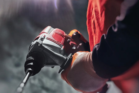 Close-up of a worker in safety vest and gloves holding a red electric drill with a drill bit for...