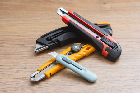 Set of utility knives and box cutters of different colors lying on a wooden surface. Concept of construction tools, office equipment, and DIY crafts