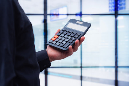 Hand in a business suit holding a black calculator against a blurred background of a modern office or corporate interior. Concept of financial management, accounting and business calculationsの写真素材