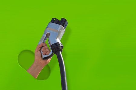Human hand holds an electric vehicle charging plug against a vibrant green background. Concept for clean energy, eco-friendly transport and electric mobility and sustainabilityの写真素材
