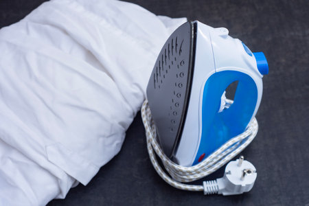 Modern electric iron with blue handle and folded white shirt on a table. Household appliance for laundry, ironing and daily clothing care at homeの写真素材