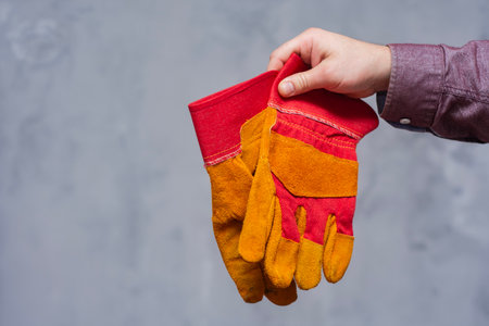 Close-up of man hand holding red and yellow protective work gloves on a neutral gray background....