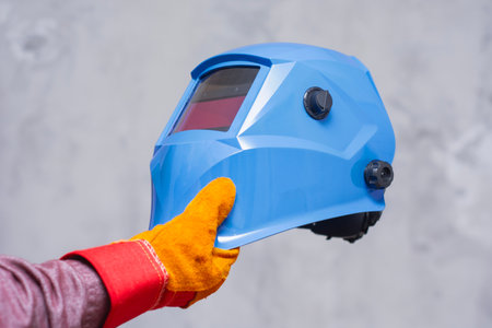 Worker hand in protective gloves holding blue welding helmet on gray concrete background. Concept of...