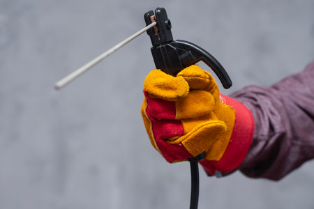 Close-up of a worker hand wearing a red and yellow protective glove holding a welding torch. The image represents industrial work, welding process and safety equipmentの写真素材