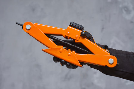 Close-up of a mechanic wearing black protective gloves holding an orange car jack tool. Concept of...