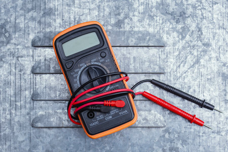 Orange digital multimeter with red test probe leads on textured gray metal background. Professional electrical measuring instrument for voltage and electrical workの写真素材