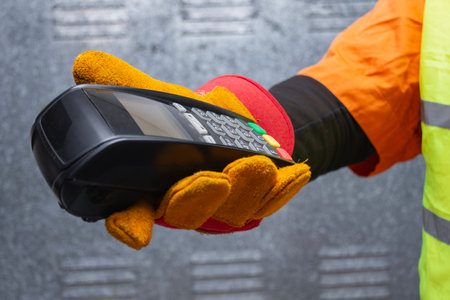 Construction worker wearing protective gloves and reflective workwear holding a wireless payment...