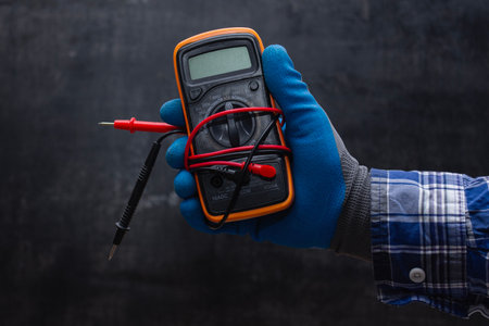 Electrician wearing a blue protective glove holds a digital multimeter with voltage probes against a dark workshop background. Tool for electrical installation work and DIY repairsの写真素材