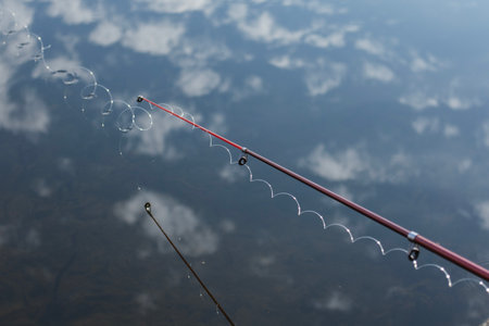 Red fishing rod tip curved with water droplets over calm blue lake surface reflecting white clouds. Concept for fishing moment with tackle equipment during outdoor recreationalの写真素材