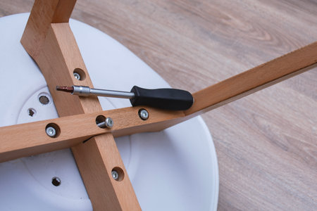 Close-up of furniture assembly process showing screwdriver, metal screws, and wooden chair parts with white metal bracket on laminate floorの写真素材