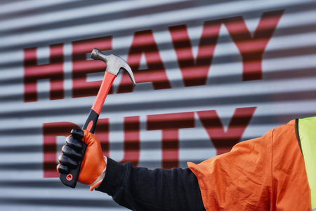 Construction worker wearing protective gloves holds a hammer against a metal wall with the words Heavy Duty written on it. Conceptual images of renovation, construction industry, and heavy laborの写真素材