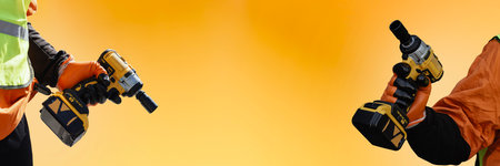 Two construction workers holding cordless drills on a bright yellow background. Concept for teamwork, modern construction tools, efficiency and industrial powerの写真素材