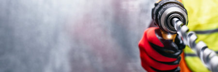 Man worker hand in protective gloves holding a power drill with a metal drill bit. Industrial background with copy space for construction, repair, and professional tool useの写真素材