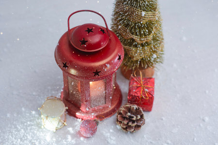 Red Christmas lantern glows beside a festive tree, ornaments, pinecone and gifts in a snowy winter setting. Holiday composition for Christmas, celebration and seasonal decorationの写真素材