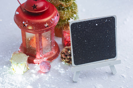 Winter Christmas mockup scene with a blank chalkboard on stand, red lantern, small Christmas tree and festive decorations on snow background. Template for holiday greetings and New Year messagesの写真素材