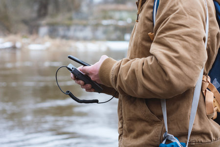 Close up of man charging smartphone with portable power bank near river. Outdoor lifestyle concept, travel technology, mobile charging, hiking equipment, connectivity in nature