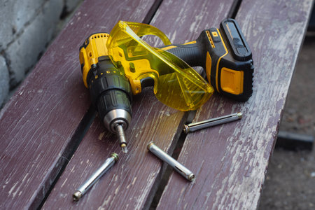 Yellow cordless drill driver with battery pack lies on a wooden bench alongside transparent yellow...