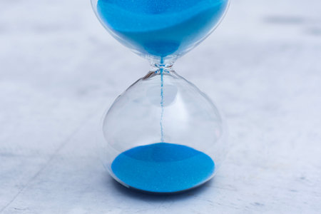 Clear glass hourglass with vivid blue sand on a light marble surface with a clean white background. Concept passing time, deadlines, urgency and limited resources in business and finance