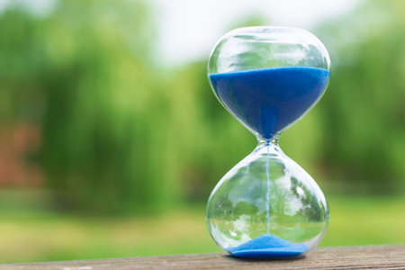 Hourglass with blue sand on wooden surface, green nature background. Time management, deadline countdown or business planning concept for corporate or marketing design