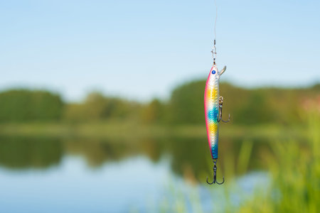 Fishing lure and colorful wobbler hanging over lake water with natural landscape background. Outdoor fishing equipment concept for angling brands and sports marketing