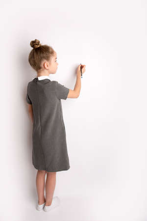 Attractive little girl in a strict school dress, stands on a white background with empty copy space, gestures with her hands, admires a happy expression on her faceの写真素材
