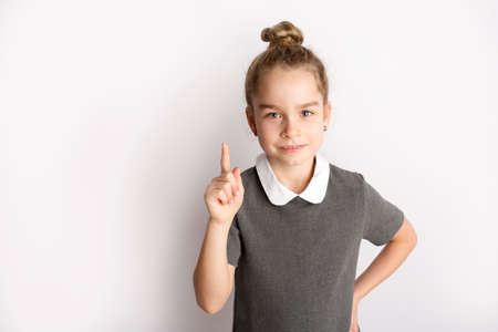 Attractive little girl in a strict school dress, stands on a white background with empty copy space, gestures with her hands, admires a happy expression on her faceの写真素材