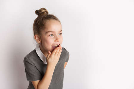 Attractive little girl in a strict school dress, stands on a white background with empty copy space, gestures with her hands, admires a happy expression on her faceの写真素材